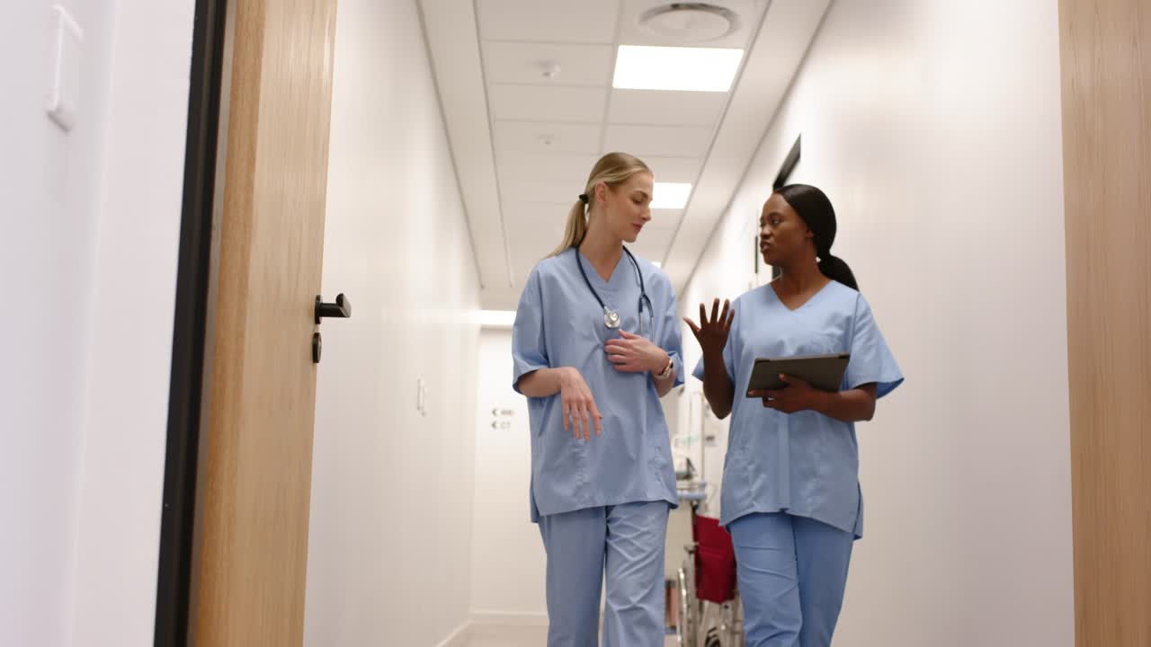 Diverse female RNs in scrubs walking hospital hall, right RN pointing tablet, planning care by cart