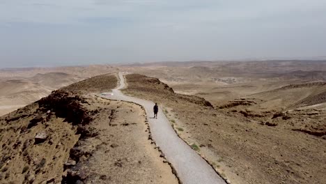 aerial-backward-slow-drone-shot-of-a-single-person-walking-on-a-mountain-trail-in-the-middle-of-an-empty-desert-hill-on-a-sunny-day