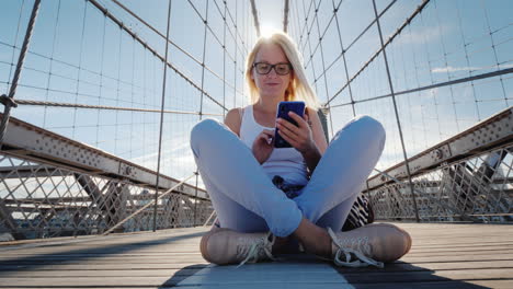 Una-Mujer-Sentada-En-El-Puente-De-Brooklyn-Usa-Un-Teléfono-Inteligente-Viaje-A-Nueva-York