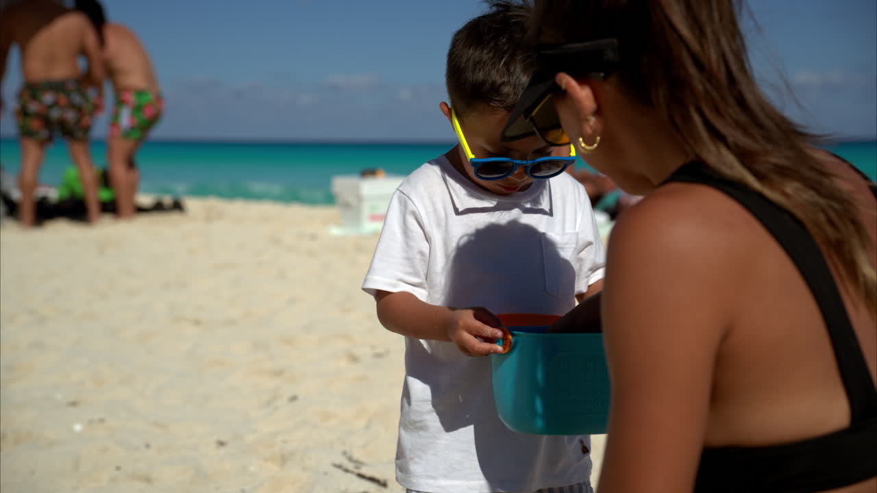 un lindo niño bruneta latino mexicano con gafas de sol y una camiseta blanca buscando conchas con su madre en una playa de cancún, méxico
