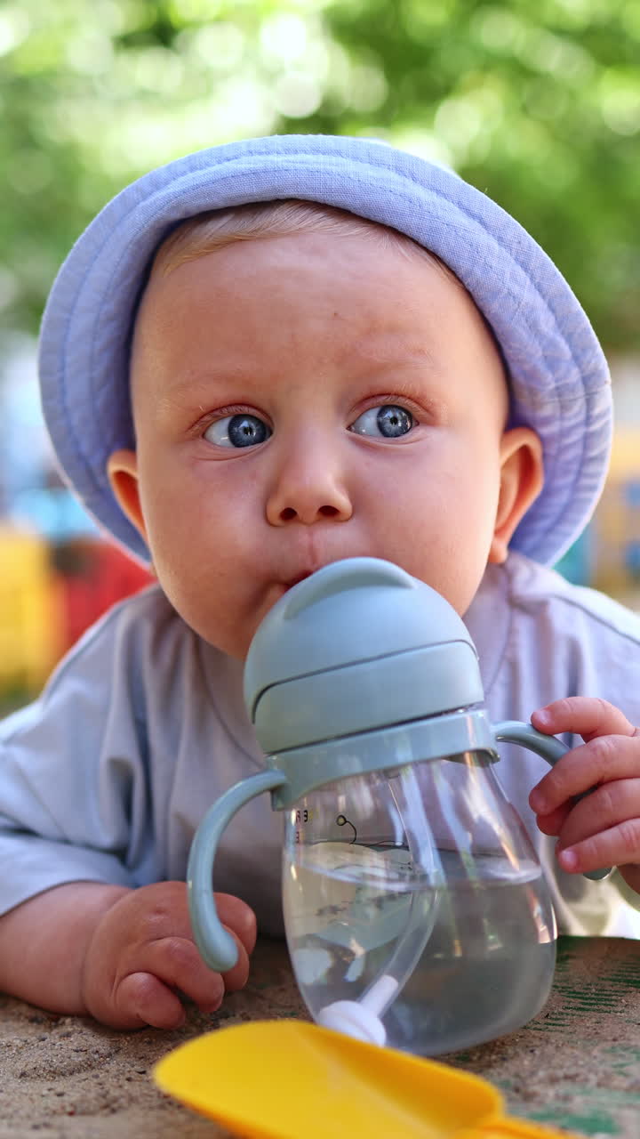 A baby boy in a linen panama on his head drinks water from a drinking bowl with a tube. Children's dishes for drinking water