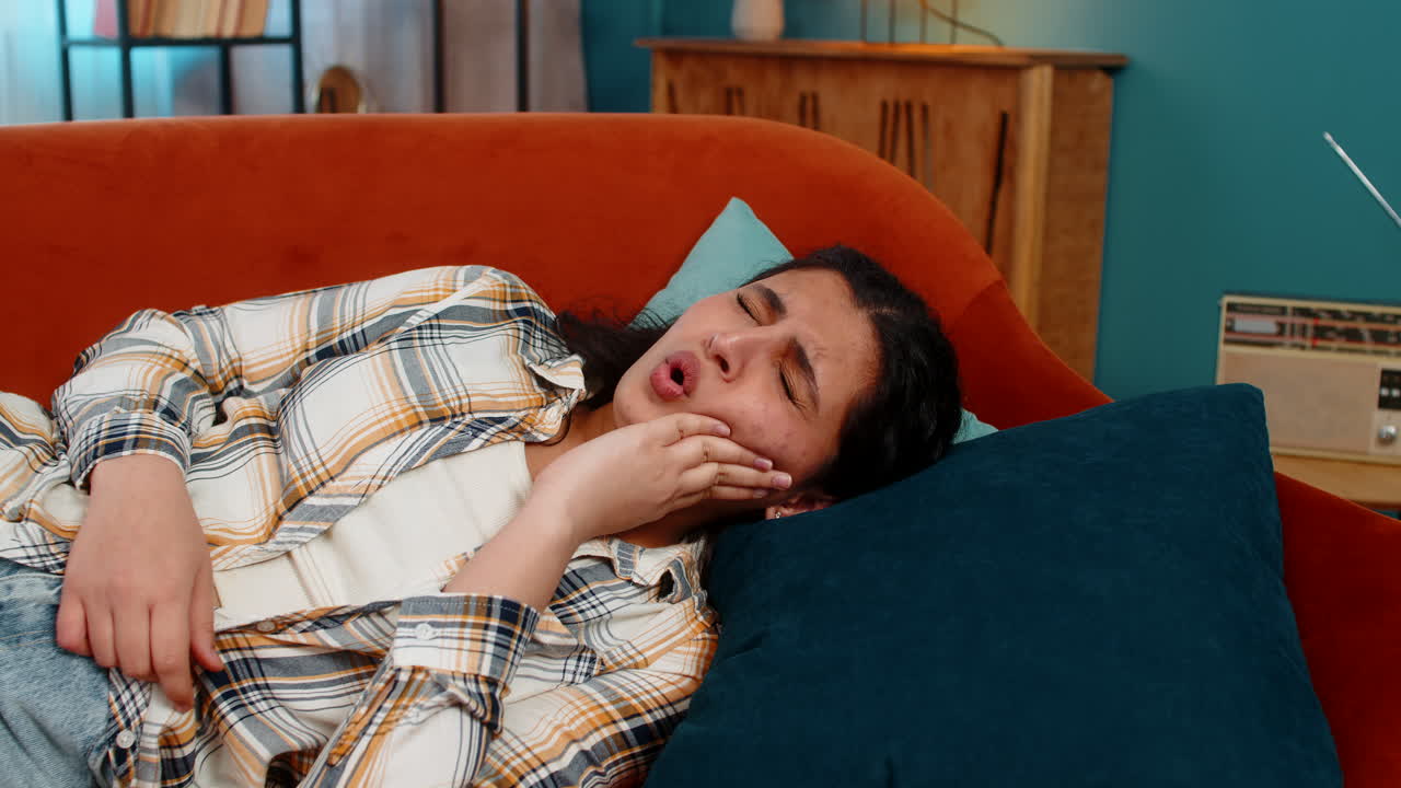 Indian woman holding cheek lying on sofa experiencing severe toothache pain discomfort distressed