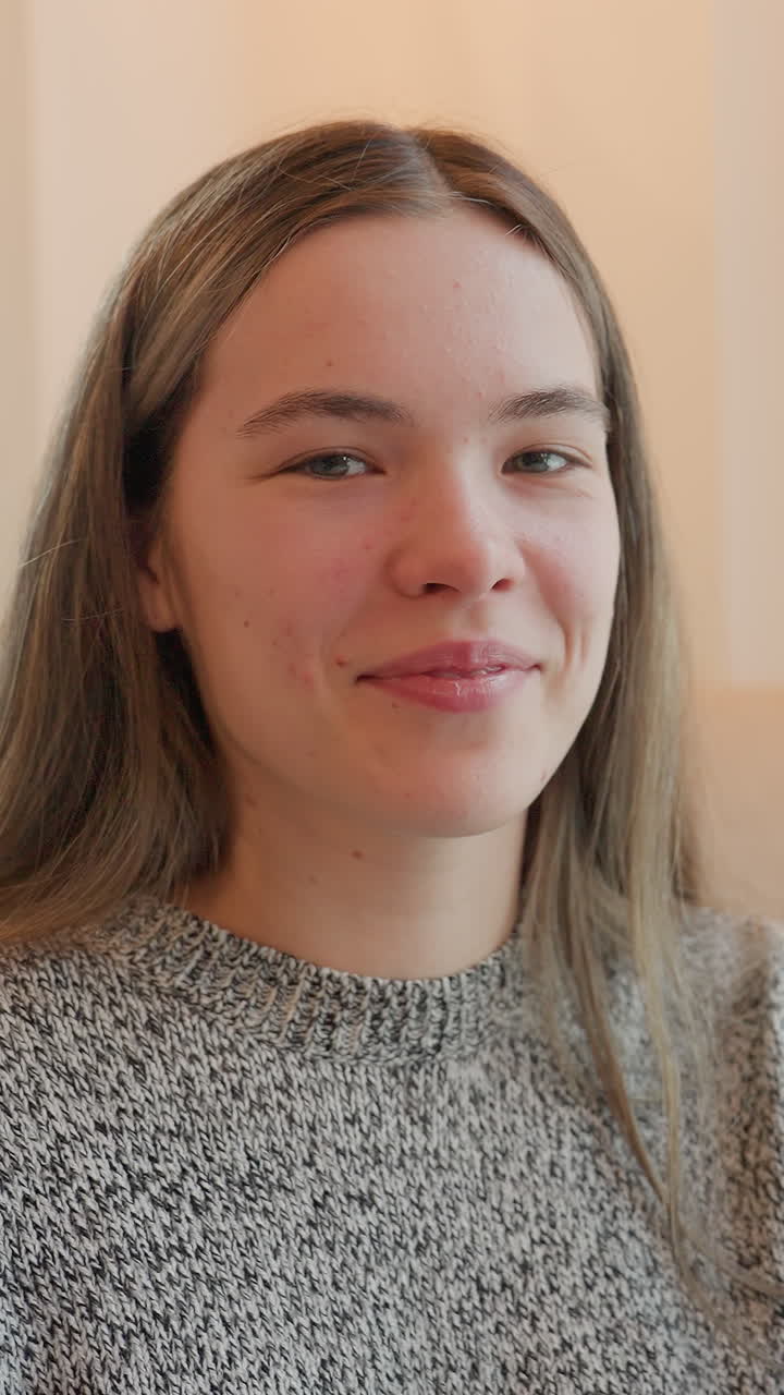 Retrato de una mujer joven con luz natural, primer plano en interiores con bokeh suave junto a la ventana, sonrisa relajada, pecas sutiles, pelo largo, jersey texturizado, iluminación ambiental cálida que sugiere una escena de estilo de vida.