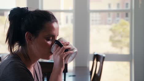 Toma-Estática-Del-Lado-Medio-De-Una-Hermosa-Mujer-Caucásica-Milenaria-Que-Desliza-Lentamente-Una-Taza-De-Café-Por-La-Mañana-Mientras-Está-Sentada-En-Su-Cocina-Y-Comedor