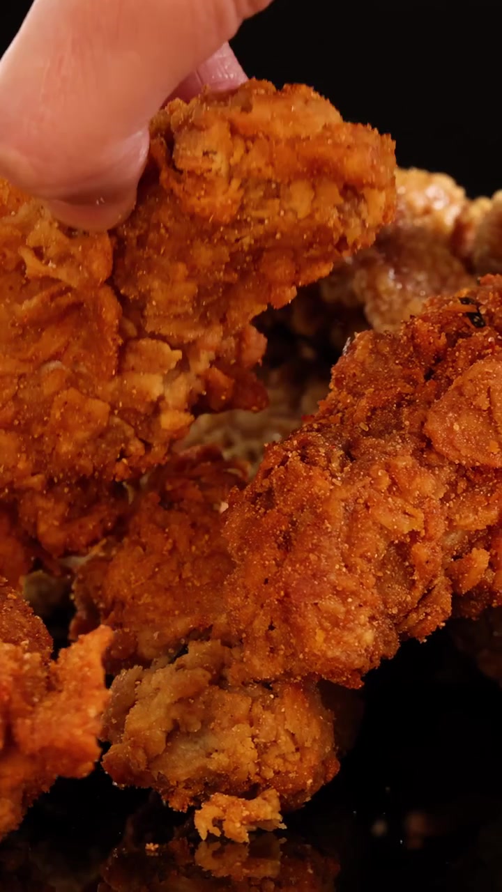 Una mano raccoglie ali e cosce di pollo fritte croccanti assortite da un mucchio sotto una calda illuminazione da studio su uno sfondo nero, con un movimento fluido della telecamera