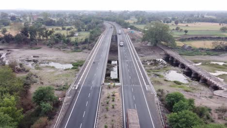 Dron-Bajando-Para-Establecer-Una-Carretera-Nacional-De-La-India-Bien-Mantenida-De-Aspecto-Genérico-Con-Un-Paisaje-Rural-De-Tierras-De-Cultivo-Y-Un-Río-Con-Camiones-Pesados-De-Transporte-De-Mercancías-Comerciales-Conduciendo-Por