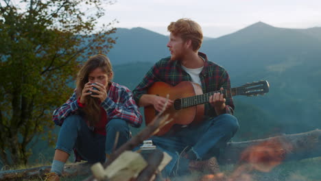 young tourists enjoy campfire in mountains. hipsters couple play music on nature