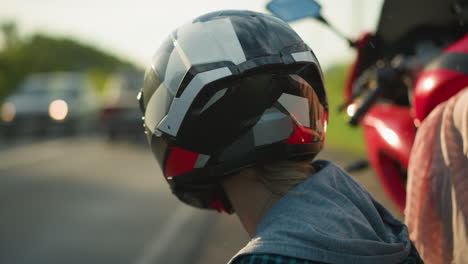 primer plano de una mujer con casco, observando los coches que pasan por una carretera, una motocicleta roja borrosa y la ropa de alguien son visibles en el fondo