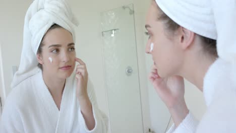 Caucasian-woman-in-bathrobe-with-towel-on-head-applying-beauty-face-mask-in-bathroom,-slow-motion