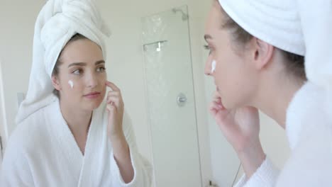 caucasian woman in bathrobe with towel on head applying beauty face mask in bathroom, slow motion