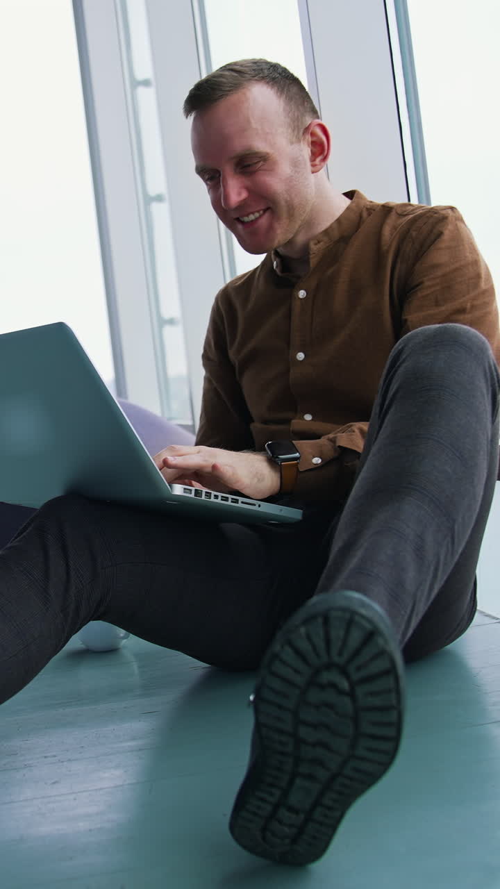 Smiling young man in modern office. Happy freelancer sitting on a floor and working on a laptop on window city view background. Vertical video