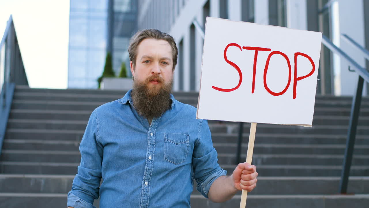 Portrait of bearded caucasian man holding Stop" signboard and looking at camera in the street"