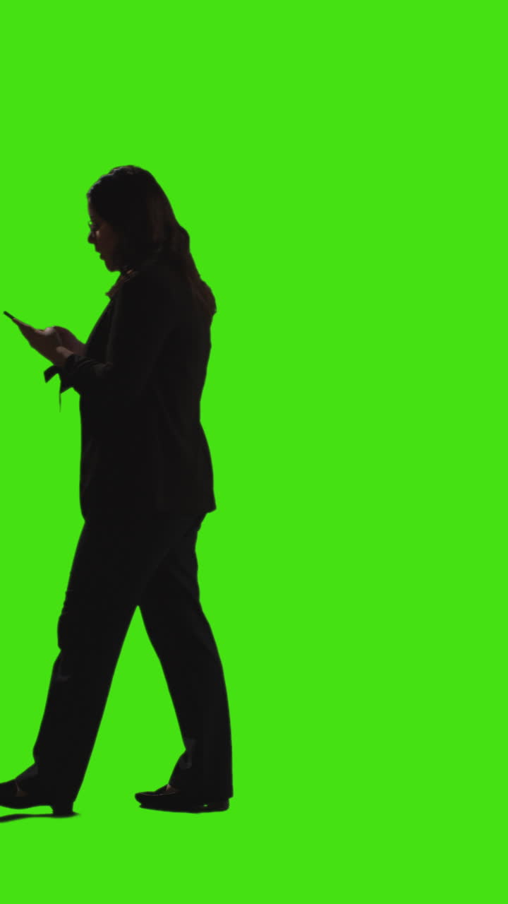 vista lateral vertical de video de una mujer de negocios enviando mensajes de texto en un teléfono móvil caminando a través del cuadro contra la pantalla verde