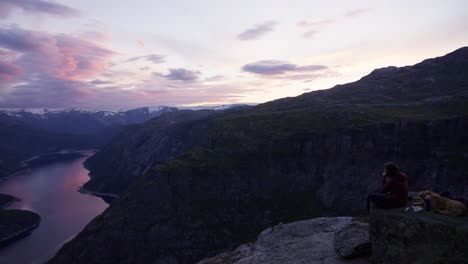 woman with golden retriever sit on a cliff at sunset, overlooking a norwegian fjord and mountains, trolltunga