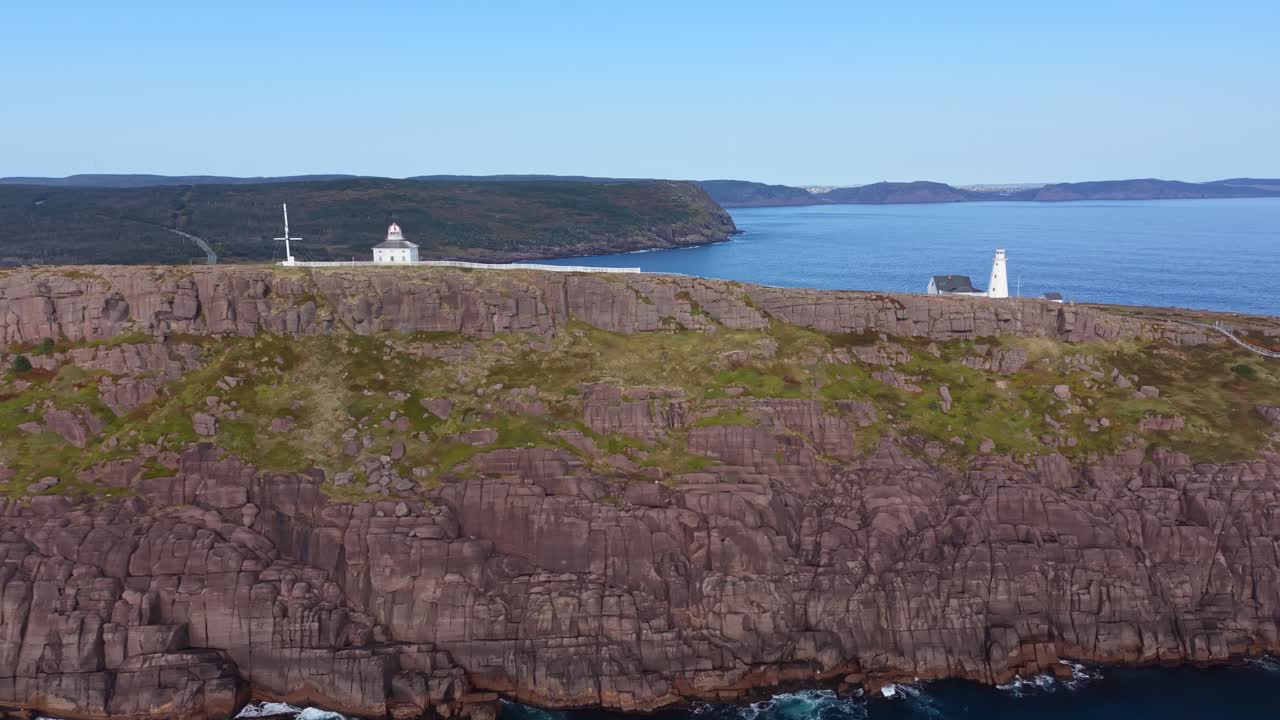 A high aerial view of Cape Spear captures twin lighthouses on a windswept plateau with Signal Hill in the background, filmed from outside the national park border