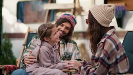 Happy-little-girl-in-a-pink-hoodie-communicates-with-her-mom,-a-brunette-girl-in-a-white-hat-and-plaid-shirt,-and-with-her-dad,-a-man-in-a-green-plaid-shirt,-during-her-vacation-and-picnic-near-a-trailer-outside-the-city-in-summer