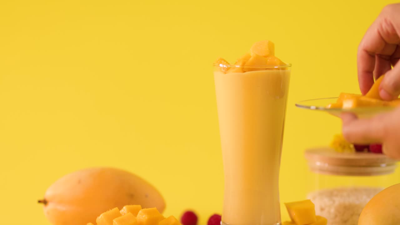 Close-up of hand adding mango pieces to tall smoothie glass on bright yellow background