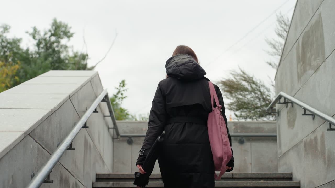 Una mujer sube unas escaleras de hormigón, una viajera con abrigo negro y un bolso rosa bajo un cielo nublado, barandilla de metal, arquitectura urbana de piedra, cámara lenta y estable desde atrás, ambiente cinemático melancólico, ideal para contenido de estilo de vida.