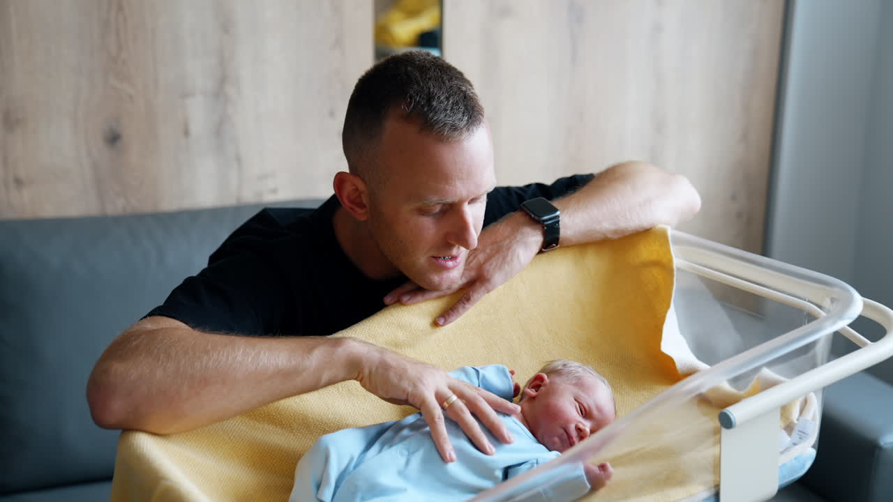Caring Caucasian dad kneeled near the baby crib. Happy parent talks to his newborn stroking his belly.