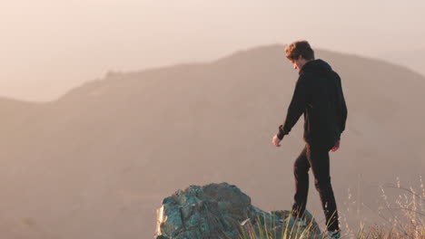 Young-adult-steps-out-onto-the-ledge-of-a-steep-mountain-overlooking-foggy-California-valley-at-sunset