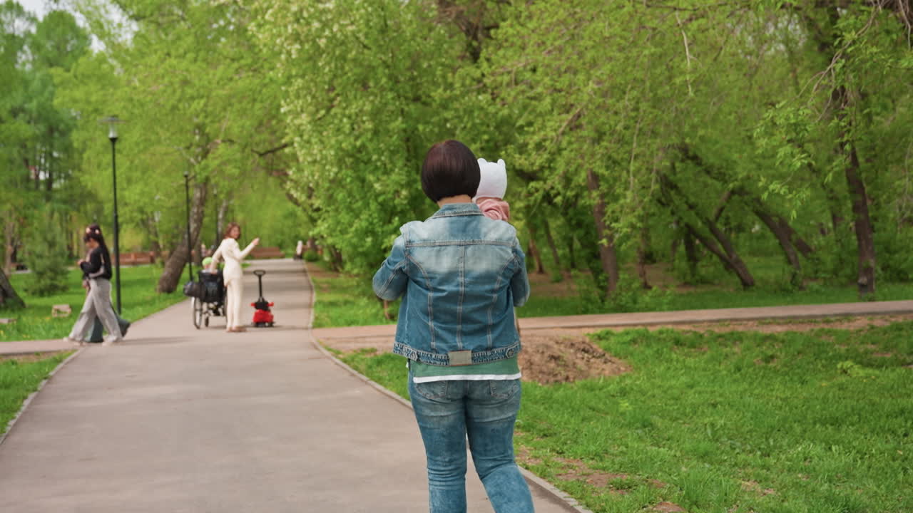 Path Approach Person Photographing Family On Walkway With Wheelchair In Frame, Casual Denim Jacket, Waving Greeting, Green Canopy, Candid Capture, Friendly Interaction, Observational Angle