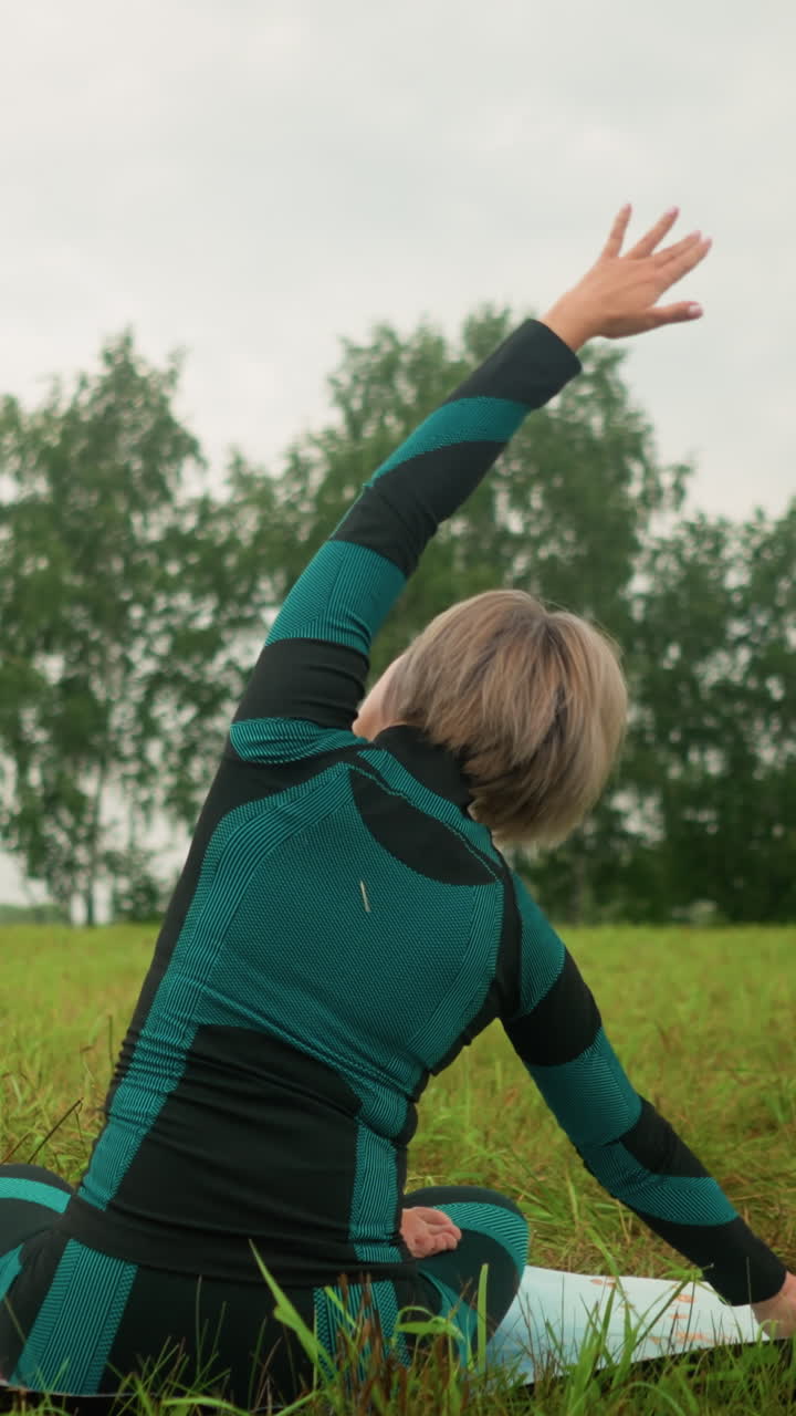 vista trasera mujer de cabello gris acostada en un lado de la alfombra de yoga practicando la postura de flexión lateral, brazos extendidos, elevándose lentamente en un campo de hierba sereno bajo un cielo nublado con árboles en la distancia