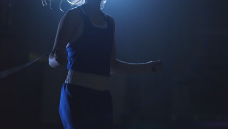 hermosa mujer boxeadora profesional en entrenamiento en el pasillo salta en una cuerda a la luz de la contra en una habitación oscura con ropa azul