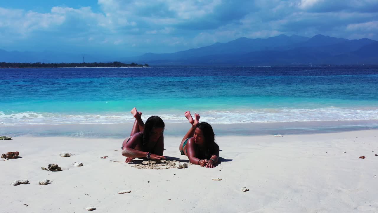 Two girls tanning lying on white sand of exotic beach washed by waves of blue azure sea on a cloudy day in Ko Samui, Thailand