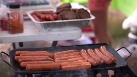 hot dogs meat beef pork and veggie sausages vegetarian plant based cooking on cast iron skillet at campsite with people walking around in background, wide camera shot