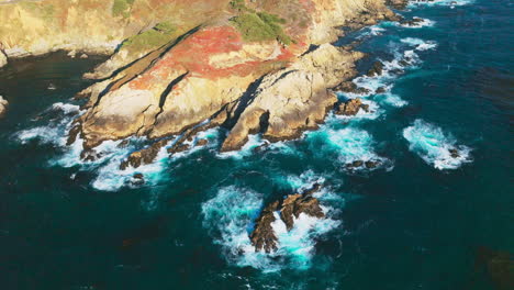 pacific ocean meeting the rocky shore of california. foamy white waves crushing at rugged bare mountains on sunny day. top view.