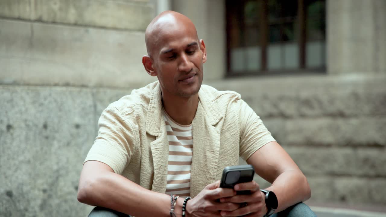 Un uomo afroamericano sulla trentina riceve un messaggio, sorride e scorre il telefono mentre è seduto su una panchina di pietra.