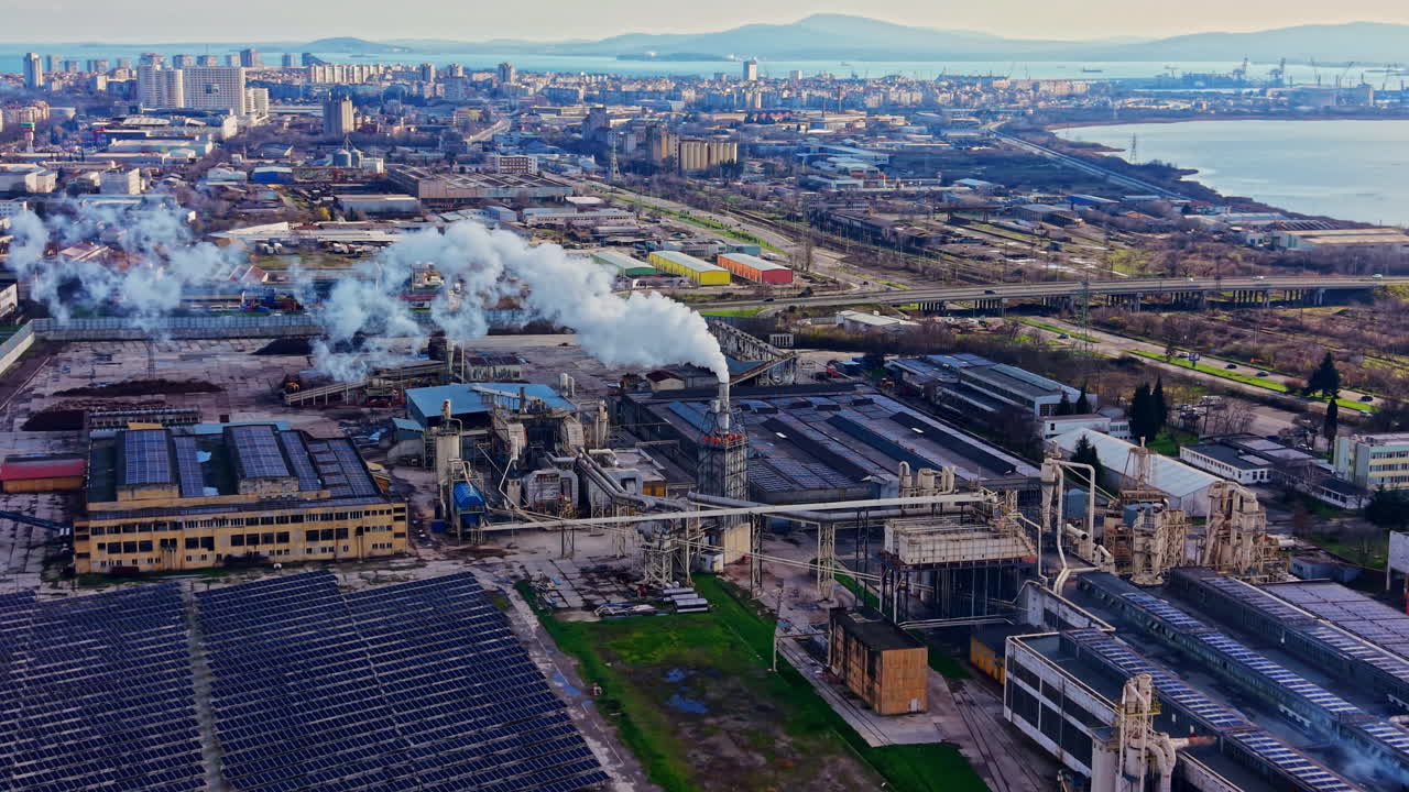 Sito industriale con fabbriche e fumo che si innalza sopra gli edifici in una città vicino a uno specchio d'acqua durante le ore diurne.