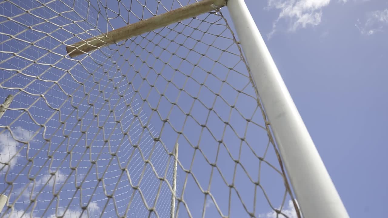 White soccer goal net gently swaying in warm summer breeze, casting subtle shadows against bright blue sky with soft cloud formations