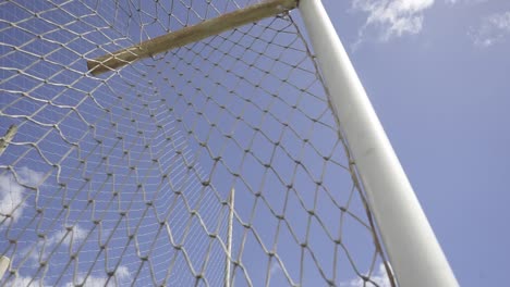 white soccer goal net gently swaying in warm summer breeze, casting subtle shadows against bright blue sky with soft cloud formations