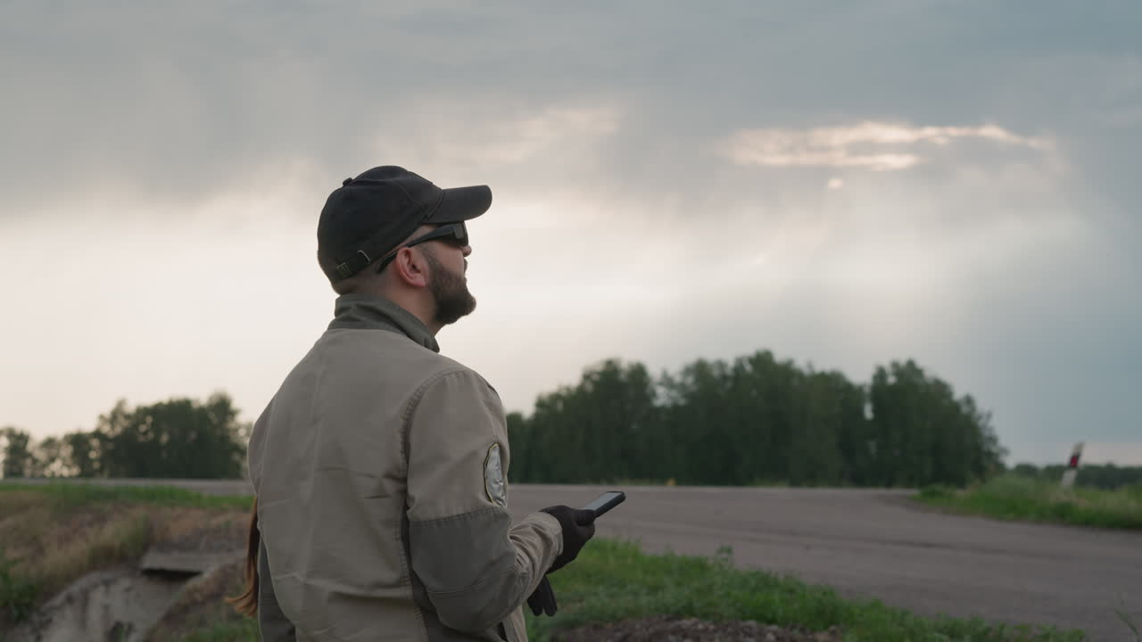 close up side view of field worker holding smartphone device while monitoring weather under cloudy sky beside rural road in overcast conditions among vegetation