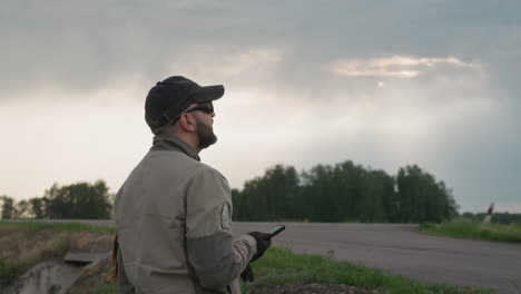 close up side view of field worker holding smartphone device while monitoring weather under cloudy sky beside rural road in overcast conditions among vegetation