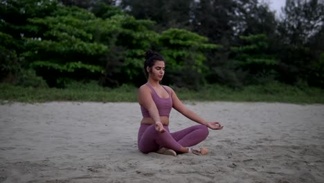 Focused-woman-in-lotus-pose-practices-yoga-on-beach,-harmonizing-body-and-mind-amidst-serene-natural-surroundings