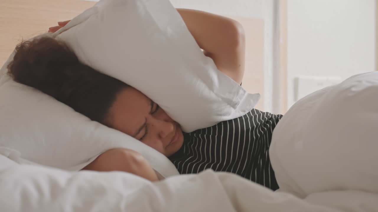Turis hotel menutupi wajahnya dengan bantal di atas selimut putih lembut, kepala ranjang kayu membingkai pemandangan, napas pelan dan ekspresi tersembunyi menunjukkan kelelahan mendalam setelah hari perjalanan yang panjang dan upaya untuk menghalau gangguan.
