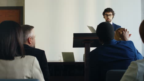 orador de negocios caucásico en un podio en una conferencia, sosteniendo documentos e interactuando con personas en el salón durante su discurso