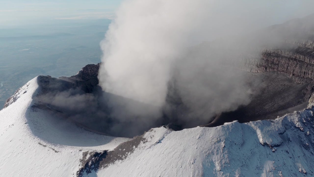 Oblique drone view across the snow-covered crater rim of Popocatepetl volcano in Mexico, with a dense white plume and distant landscape beyond.