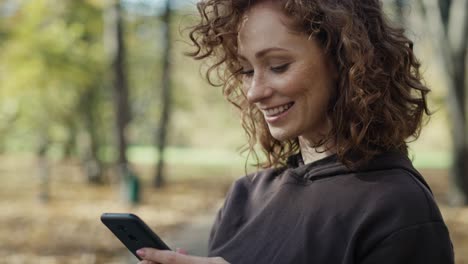 Smiling-ginger-woman-standing-in-the-park-and-using-mobile-phone