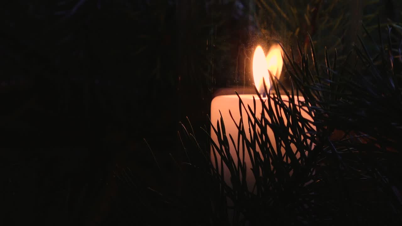 A warm candle flame flickers inside a glass lantern, softly illuminating surrounding pine needles in a dark, cozy setting