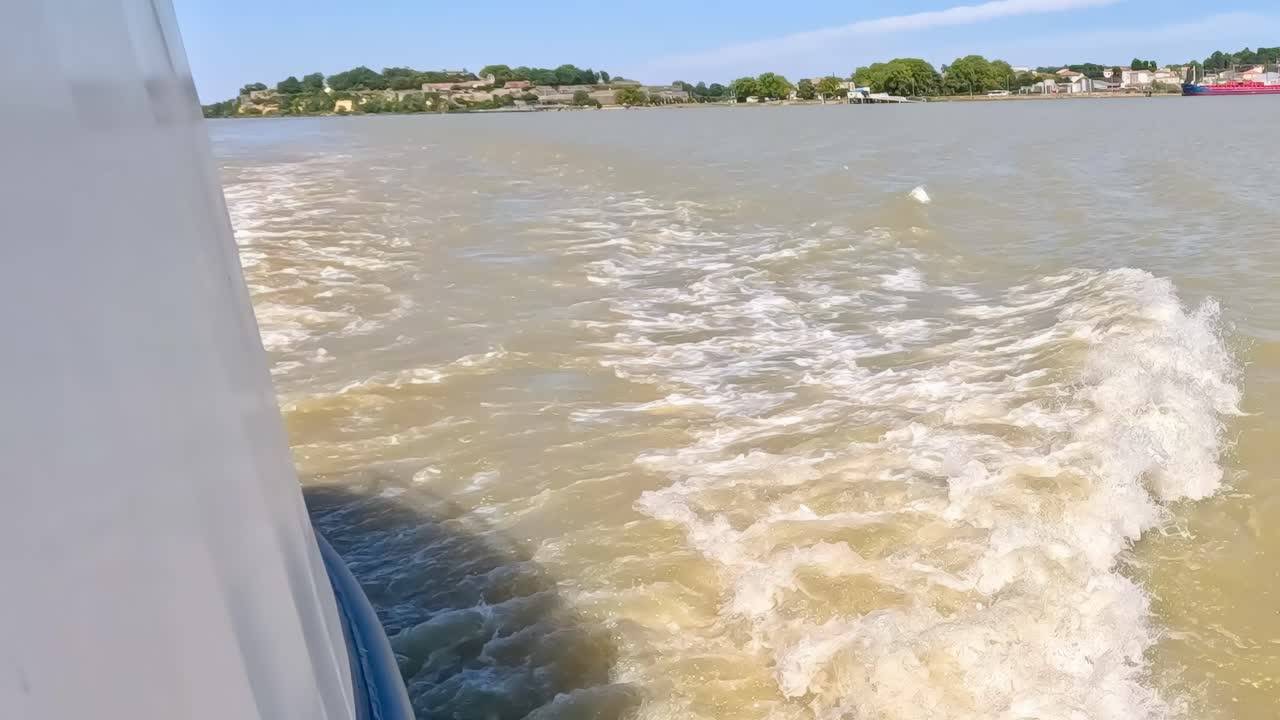 Close-up view of boat creating waves and wake on a river, with distant shoreline visible.