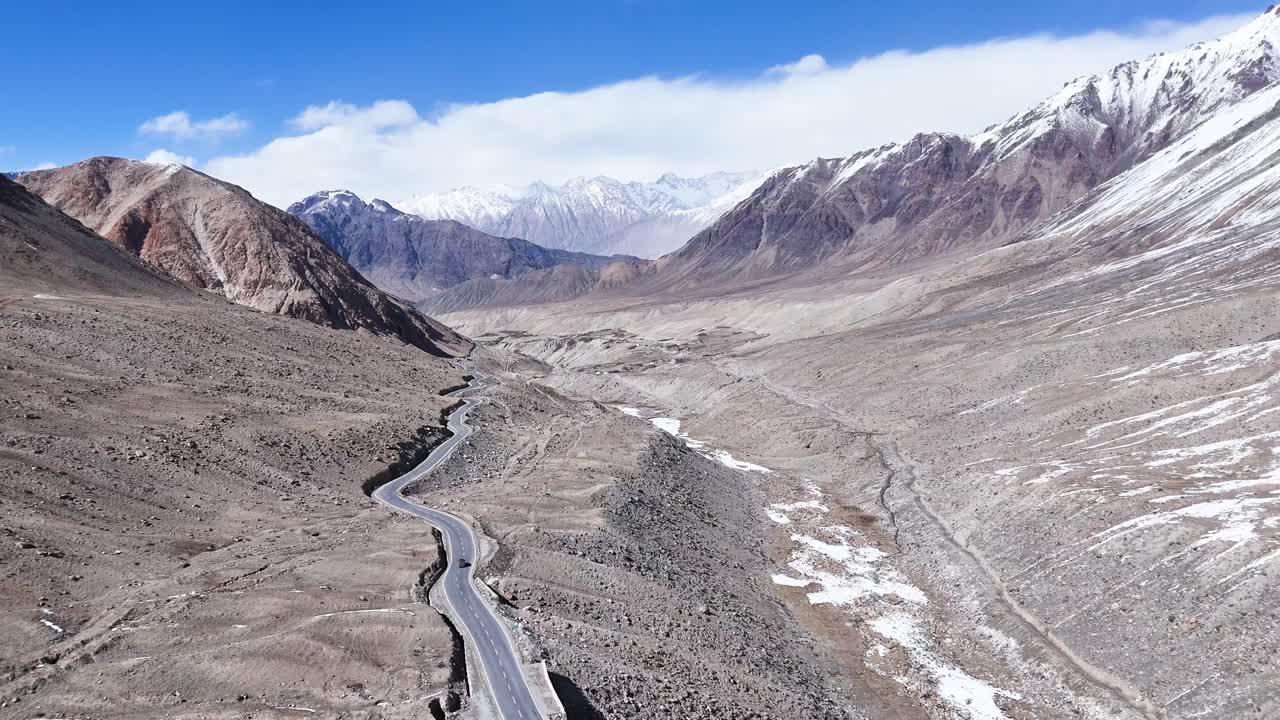 Aerial drone shot showing a vehicle driving past scattered settlements and prayer flags in Ladakh.