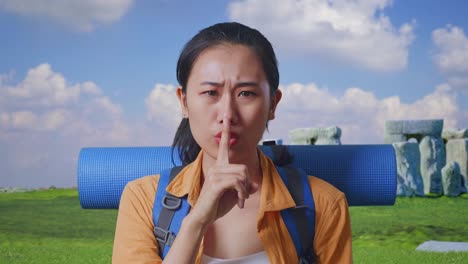close up of asian female hiker with mountaineering backpack looking at camera and making shh gesture while traveling in stonehenge