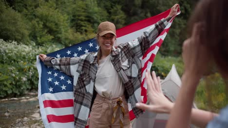 first person method a girl photographs her friend who is standing with the flag of the united states of america in a cap against the backdrop of a green forest