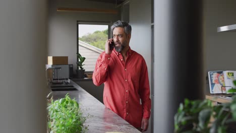 Hombre-Birracial-Con-Camisa-Roja-Hablando-Por-Teléfono-Inteligente-Solo-En-La-Cocina