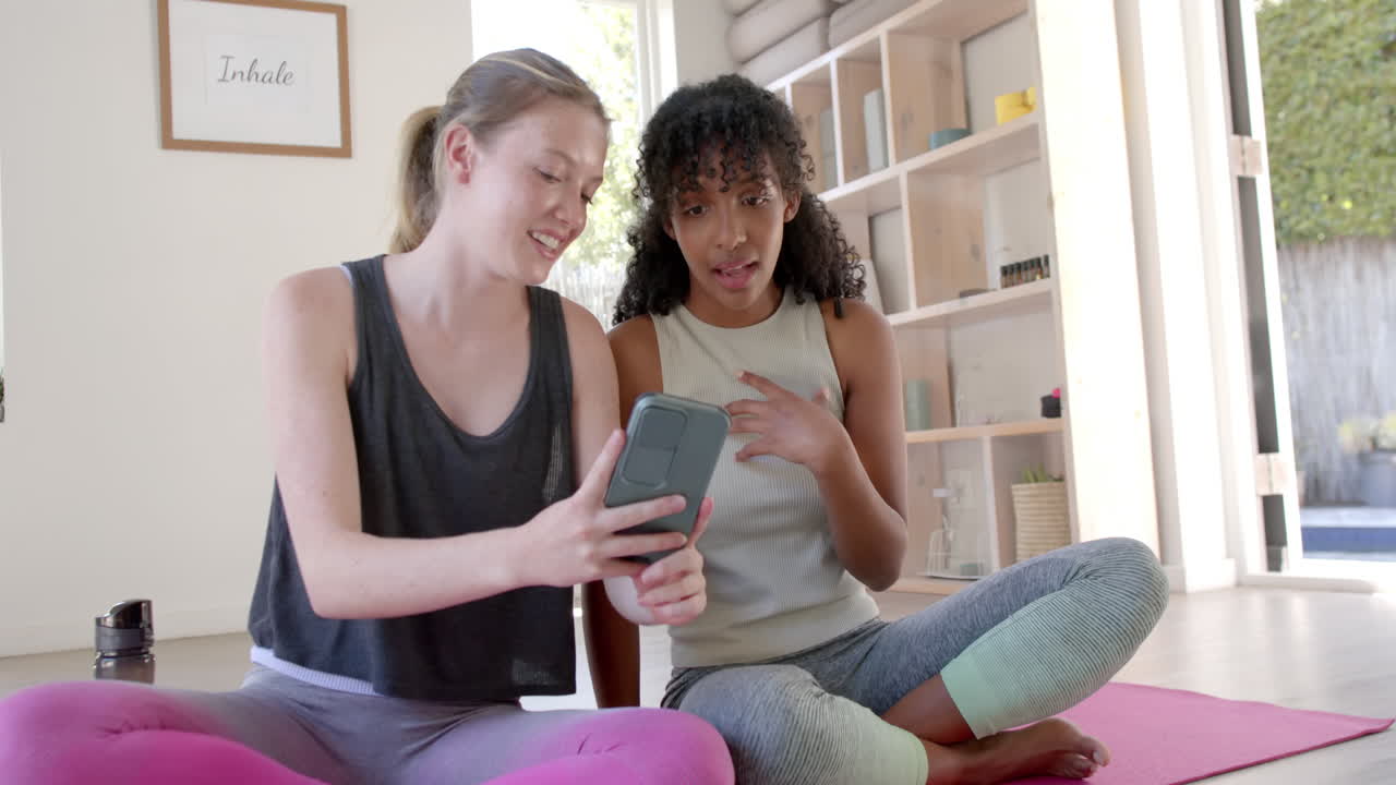 mujeres felices y diversas sentadas en esteras de yoga y usando teléfonos inteligentes, en cámara lenta