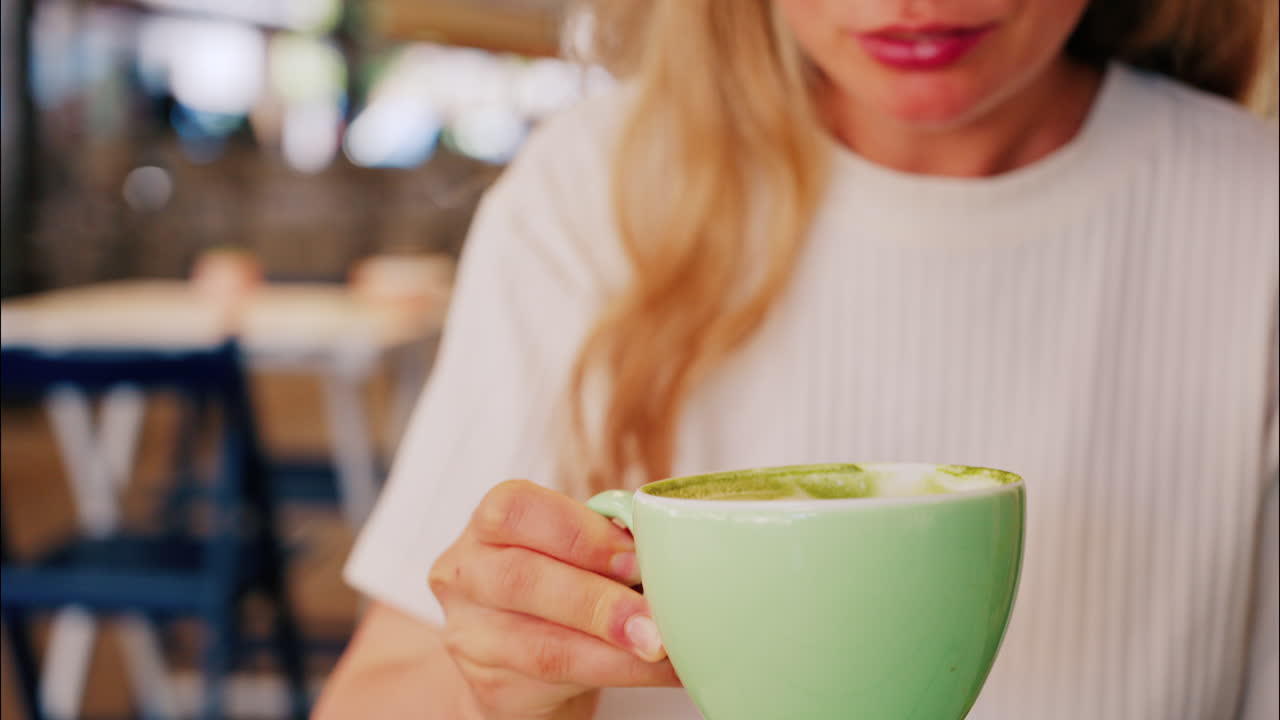 Close up of a blonde woman in a white T-shirt drinking a matcha latte out of a green cup at a terrace