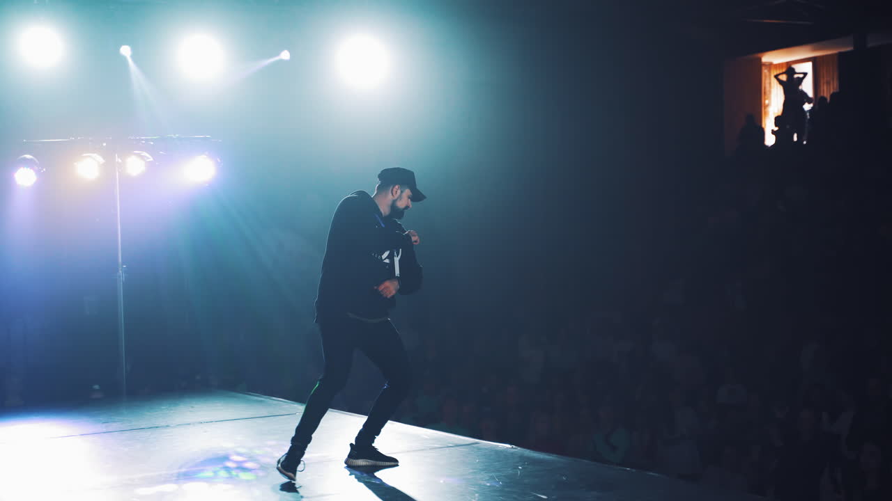 Modern dance class or performance in blur dark auditorium. Bearded man in black sport clothes entertaints the audience with his hip-hop dance.